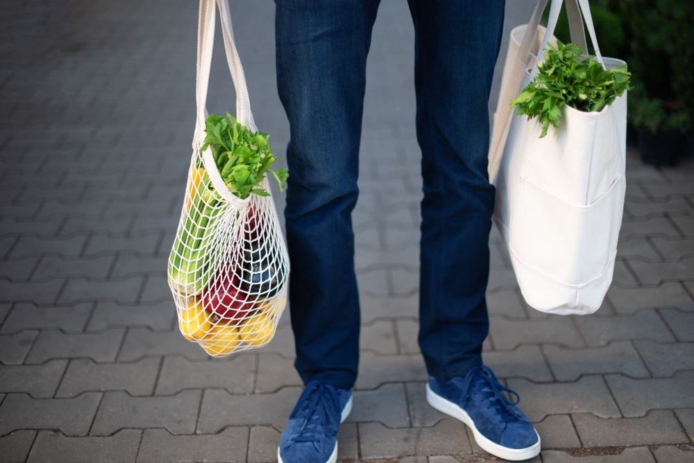 Person holding two reusable grocery bags filled with fresh produce while standing on a sidewalk